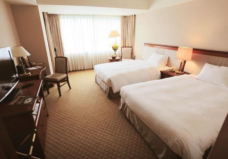 Evergreen Laurel Hotel - Taichung Eksteriør bilde The photo shows a hotel room featuring two twin beds with white bedding. There's a chair positioned near one of the beds and a desk with a computer setup against one wall. Large windows allow natural light to enter the room, and light-colored curtain
