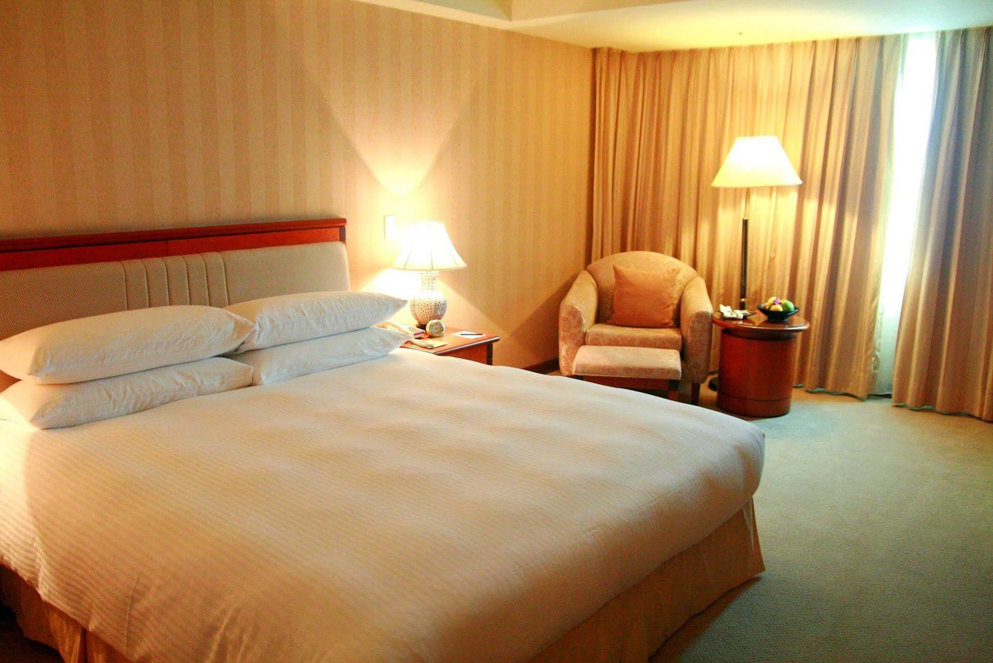 Evergreen Laurel Hotel - Taichung Eksteriør bilde The photo shows a hotel room containing a large bed with white linens, flanked by two bedside lamps. To one side, there is a cozy armchair and a small side table with a fruit bowl or snacks. The walls are decorated in a soft, neutral color, and there