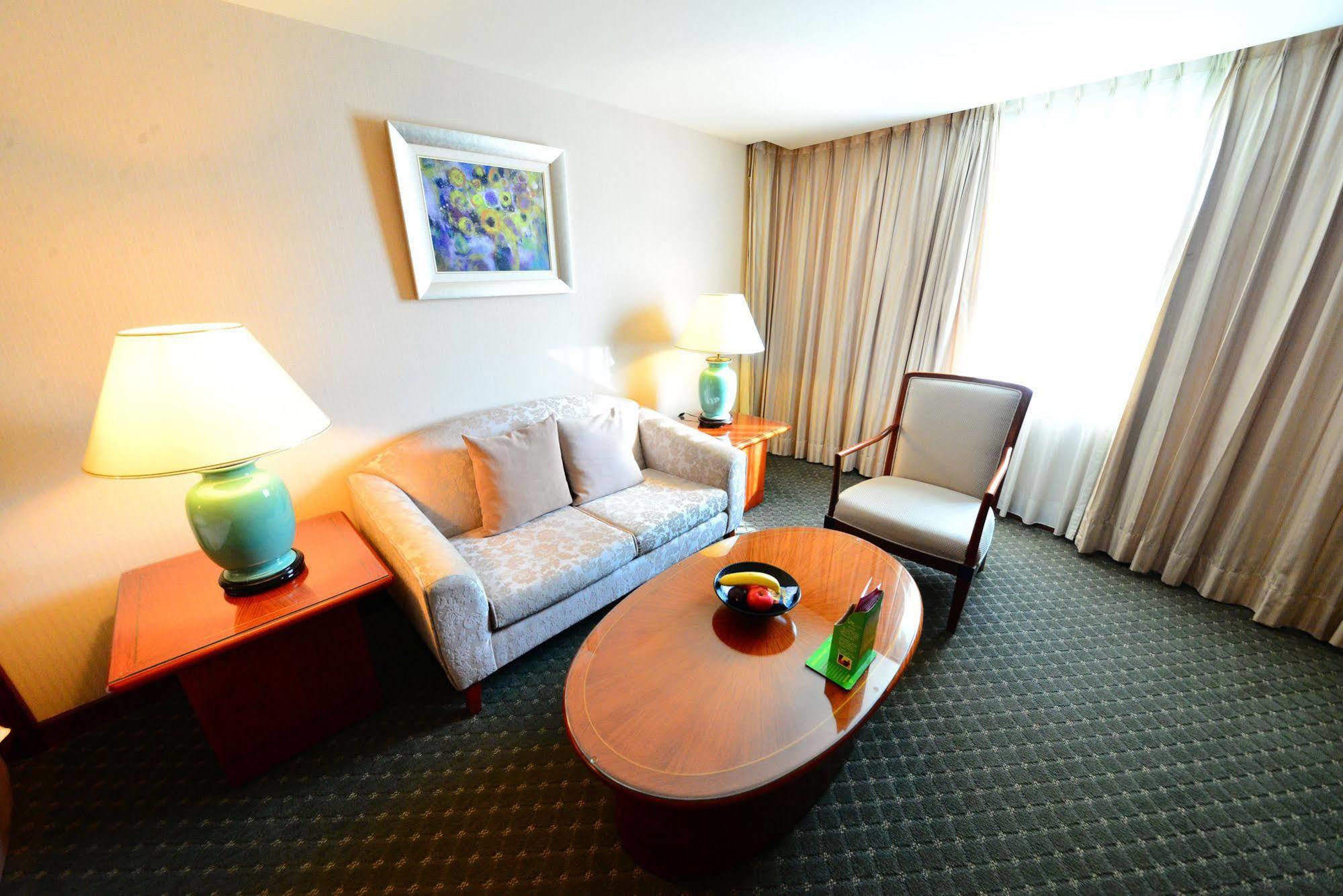 Evergreen Laurel Hotel - Taichung Eksteriør bilde The photo shows a cozy living space, likely a hotel room or a lounge area. It features a light-colored sofa with cushions, a wooden coffee table in the center, and a comfortable armchair beside it. There are two lamps with turquoise bases on side tab