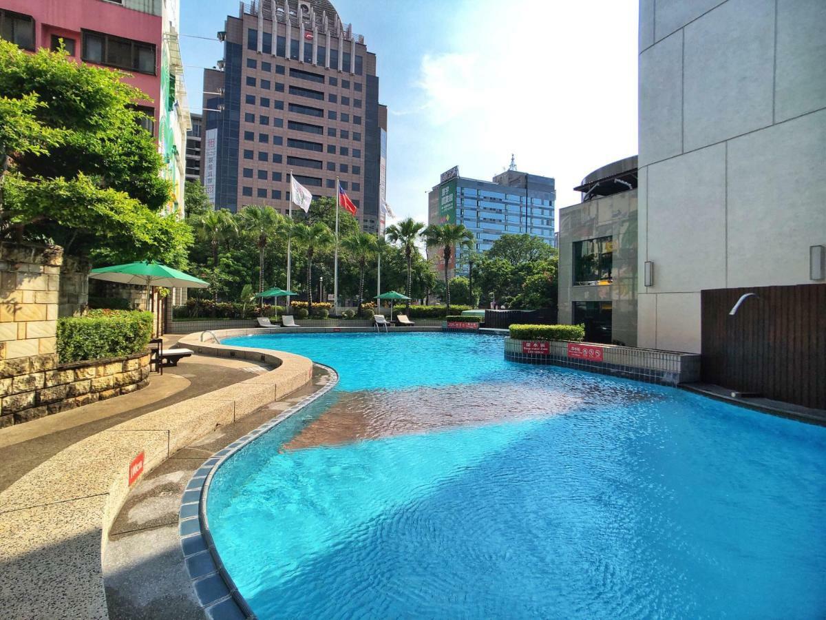 Evergreen Laurel Hotel - Taichung Eksteriør bilde The photo shows a swimming pool set in a modern urban environment. The pool has a curved edge and is surrounded by greenery, including trees and umbrellas for shade. In the background, there are several tall buildings, indicating a cityscape, with so