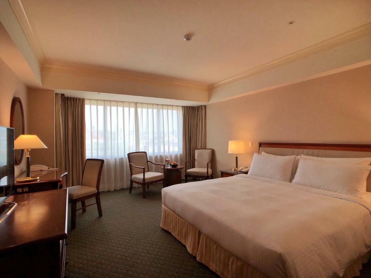 Evergreen Laurel Hotel - Taichung Eksteriør bilde The photo shows a hotel room featuring a large bed with white linens, a wooden bedside table with a lamp, and a flat-screen television. There are two chairs with a small table next to them positioned by a large window, which has sheer curtains lettin