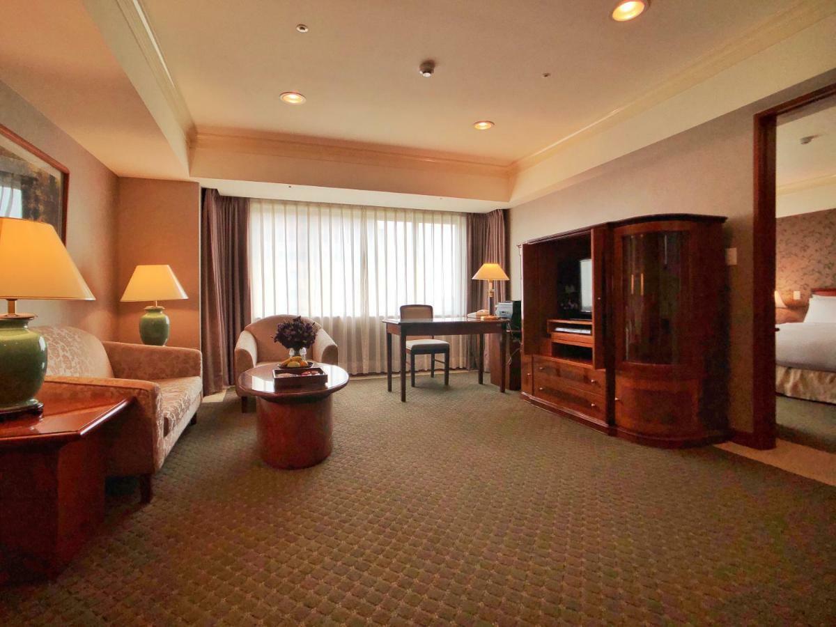Evergreen Laurel Hotel - Taichung Eksteriør bilde The photo shows a cozy hotel suite or living area. There is a sofa and an armchair arranged around a small coffee table. A desk with a chair is positioned near a large window covered with sheer curtains that allow natural light to enter the room. Two