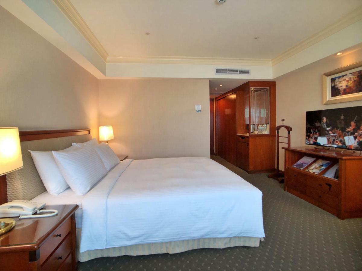 Evergreen Laurel Hotel - Taichung Eksteriør bilde The photo shows a hotel room featuring a comfortable bed with white linens and pillows, positioned against a light-colored wall. There is a wooden bedside table with a lamp and an open book next to the bed. On the opposite side, there’s a large flat-
