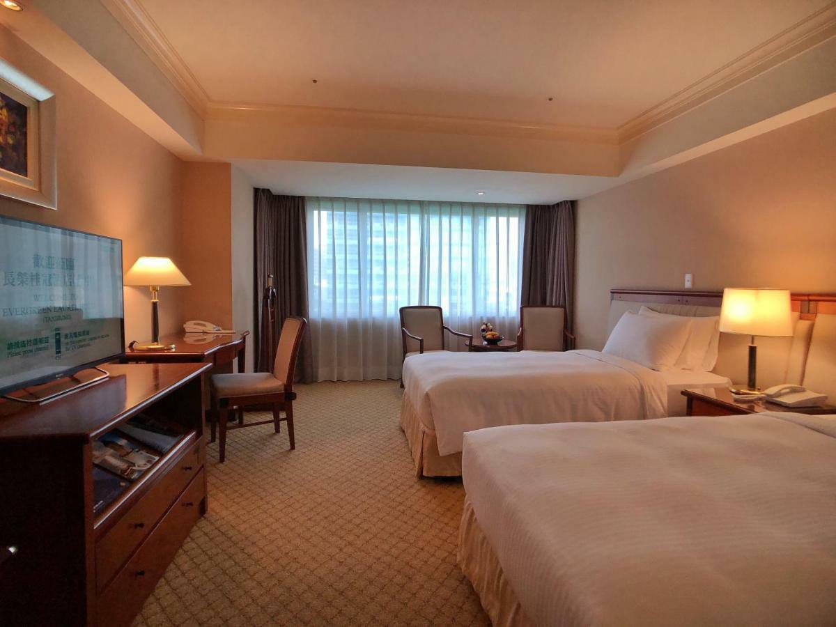 Evergreen Laurel Hotel - Taichung Eksteriør bilde The photo shows a hotel room featuring two twin beds with white bedding. There's a television mounted on a wooden dresser, and next to it, a comfortable chair and a small table are positioned. The room has large windows that allow natural light to en
