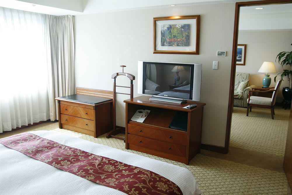 Evergreen Laurel Hotel - Taichung Eksteriør bilde The photo depicts a hotel room interior. In the foreground, there is a neatly made bed with a decorative red bedspread. To the left, a large window allows natural light to fill the room, with light-colored curtains drawn to one side. 

Next to the be