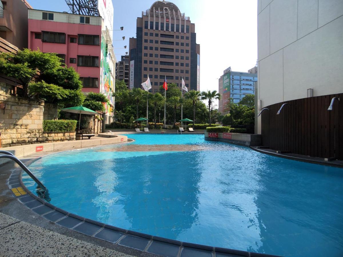 Evergreen Laurel Hotel - Taichung Eksteriør bilde The photo shows a swimming pool area set in an outdoor environment. The pool has a clear blue water and a gently curving shape. Surrounding the pool are some green trees and plants, along with a few umbrellas providing shade. In the background, there